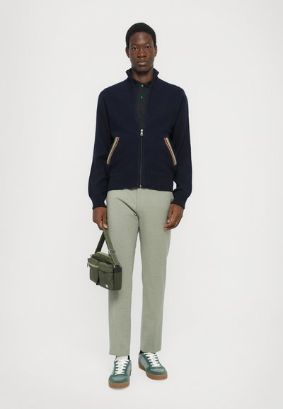 Marinblå zip-up tröja med regnbågsfärgade sidostickningar, svart polo-shirt, ljusgröna byxor, grön axelväska och teal sneakers med vita snören.