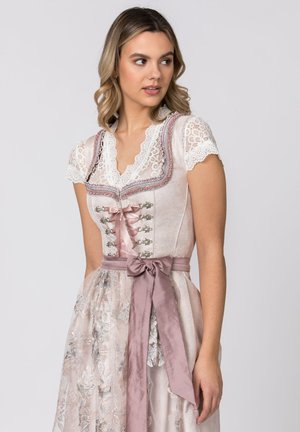 Junge Frau in einem hellrosanen Spitzen-Dirndl mit silberner floraler Schürze und einem satinartigen Band, das zu einer Schleife in der Taille gebunden ist, schaut seitlich.