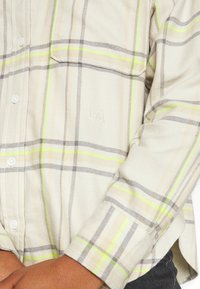 Person wearing a beige plaid shirt with yellow and gray stripes, buttoned cuff, and embroidered "L.S.S." logo near chest pocket.
