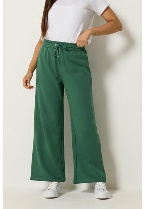 Vrouw met groene sweatpants met wijde pijpen en een trekkoord in de taille, wit T-shirt en witte sneakers, staand tegen een effen muur.