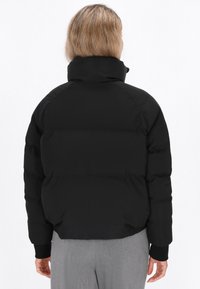 Czarna kurtka puffer z wysokim kołnierzem, o gładkiej fakturze i prążkowanych mankietach. Krój jest skrócony, podkreślający kwadratowy kształt sylwetki.