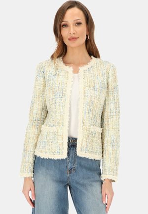 Kobieta ubrana w kremową, wielokolorową kurtkę z tweedu z frędzlami, w połączeniu z białym topem i niebieskimi dżinsami, stojąca na tle jednolitego tła.