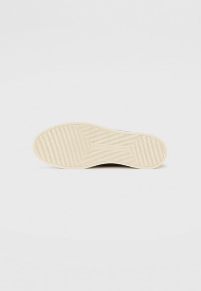 Shoe sole with a textured, patterned surface in beige, featuring a central logo and ridges for traction. Clean, minimalist design.
