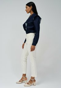 Chaqueta de mezclilla con cuello con volantes, combinada con pantalones ajustados blancos y tacones de tiras beige, mostrando una silueta limpia y ajustada.