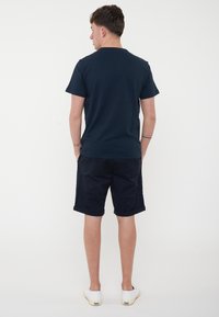 T-shirt di cotone blu navy, maniche corte; abbinato a pantaloni corti neri; scarpe slip-on bianche; in piedi con la schiena rivolta verso l'osservatore.