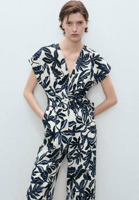 Combinaison à imprimé floral en crème, avec des motifs de feuilles noires et bleues. Manches courtes, décolleté en V et taille cintrée avec un détail de ceinture.