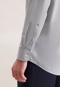 Camicia a scacchi in bianco e nero con polsino abbottonato, caratterizzata da un tessuto testurizzato e un piccolo emblema nero vicino al polsino.