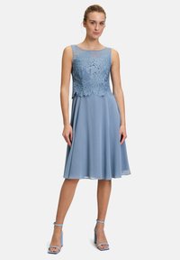 Blaues Kleid mit einem Spitzenoberteil, das Blattmuster aufweist, fließendem Rock und rundem Ausschnitt. Kombiniert mit hellblauen Riemchensandalen.