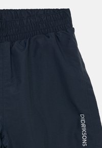 Prim-plan cu pantaloni negri închis, cu talie elastică și logo-ul "Didriksons" imprimat vertical pe piciorul drept.