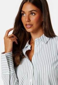 Camicia bianca con righe verdi sottili, dotata di colletto e maniche lunghe. Tessuto liscio con bottoni in vista.