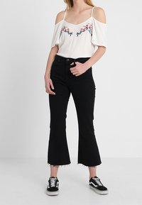 Bílá halenka s odhalenými rameny a květinovou výšivkou, kombinovaná s černými vysokým pasem a rozšířenými džíny a černými teniskami s bílými detaily.