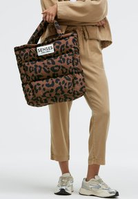 Brown leopard print tote bag with padded texture and a white tag labeled "SENSES THE LABEL." Features durable handles and a spacious design.