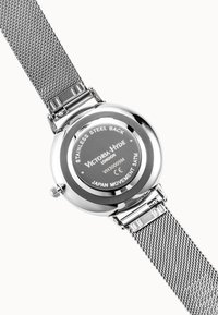 Correa de reloj de malla metálica plateada con fondo redondo de acero inoxidable grabado con la marca, modelo y detalles de resistencia al agua.