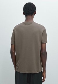 Hnědé tričko s krátkým rukávem vyrobené z měkké látky, s volným střihem, jednobarevné a s kulatým výstřihem. Bez viditelných vzorů nebo akcentů.