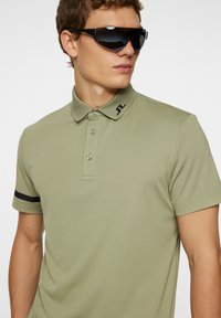 Olivgrön pikétröja med korta ärmar, tre-knappsstängning och svart accentrand. Mörka solglasögon fulländar den atletiska stilen.