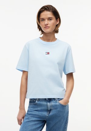 Młoda osoba z krótkimi włosami nosi jasnoniebieski t-shirt Tommy Jeans i niebieskie dżinsy, stojąc z jedną ręką w kieszeni na białym tle.