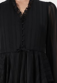 Black dress featuring a v-neckline with ruffled trim, six buttons down the front, and a tied waist. The fabric has subtle stripes and a flowing skirt.