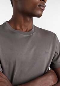 Detailní záběr osoby v šedém tričku s kulatým výstřihem, na hrudi malé vyšívané logo psa, ruce zkřížené.