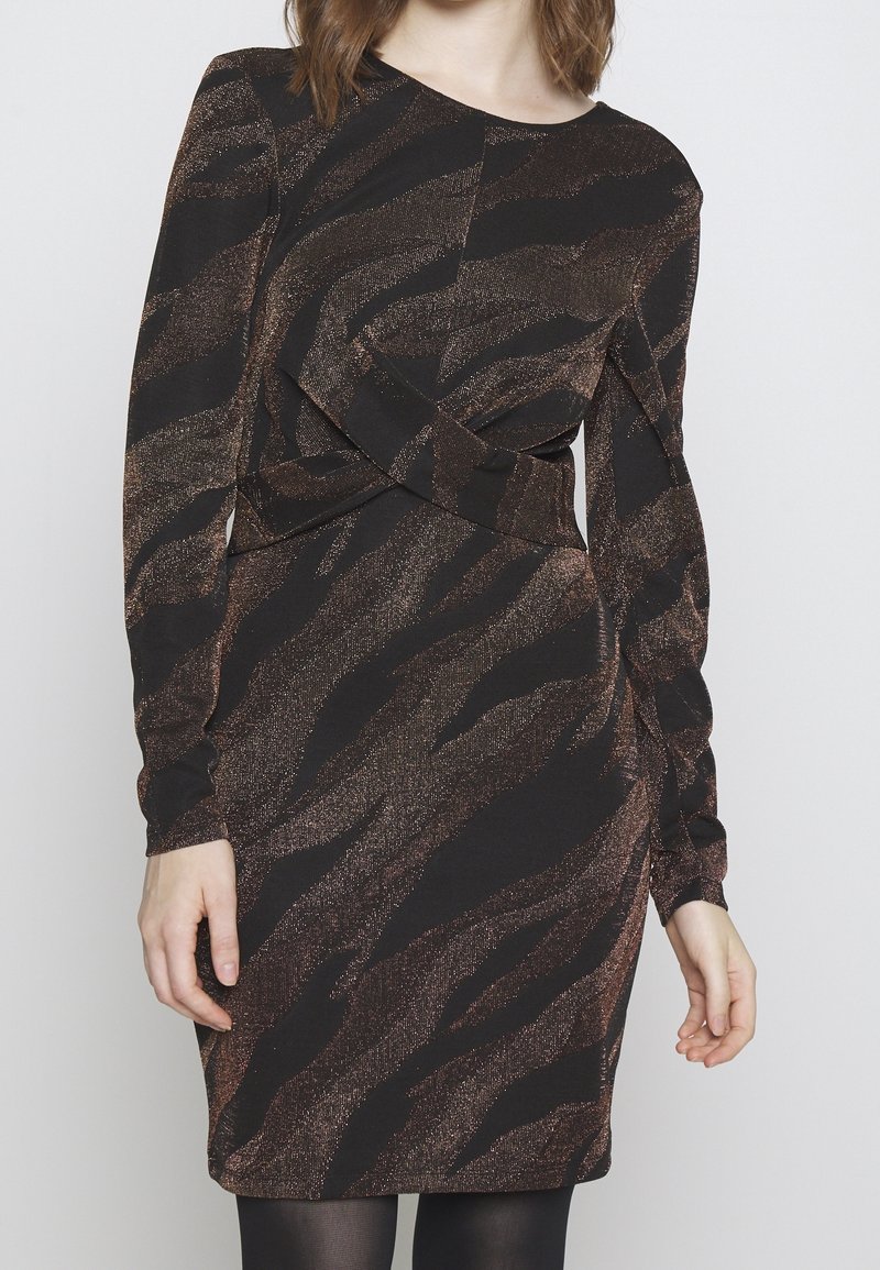 Femme portant une robe à manches longues, imprimé animal noir et marron, avec des détails asymétriques de tissu superposé à la taille.
