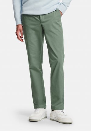 Mann trägt hellgrüne, gerade geschnittene Hosen, weiße Sneaker und einen hellblauen Pullover, eine Hand in der Tasche.