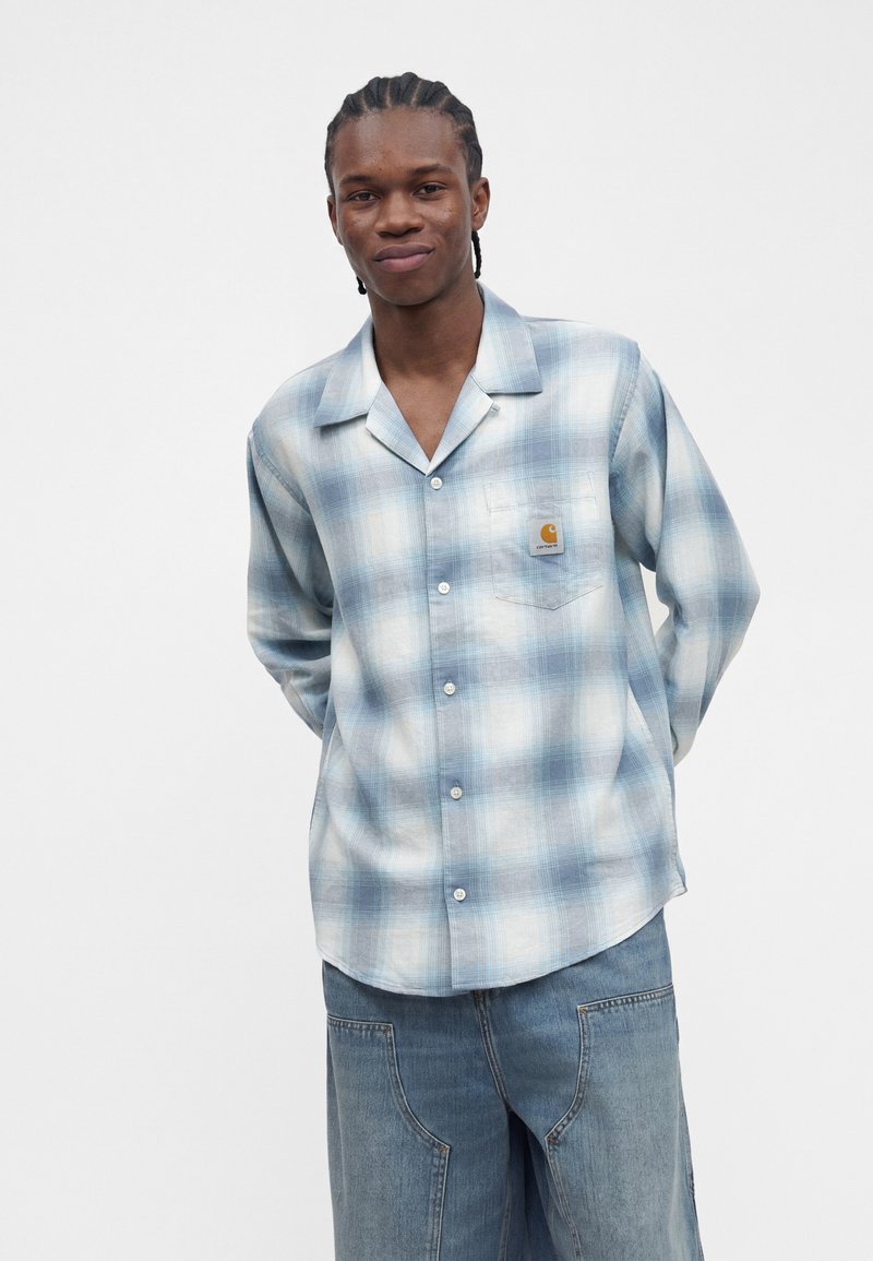 Young man with braided hair wearing a blue and white plaid button-up shirt and loose denim jeans against a plain white background.