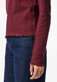 Bordo spalvos ilgomis rankovėmis viršus su ribota tekstūra, banguota apykakle ir prigludusia siluetu, derinamas su tamsios džinsinės medžiagos kelnėmis.