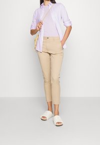 Camisa de botones color lavanda superpuesta sobre una blusa lavanda, pantalones beige ajustados y sandalias de peluche blancas. Diseño simple y colores neutros.