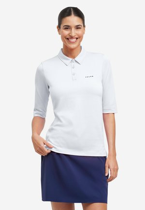 Glimlachende vrouw in een wit poloshirt met korte mouwen en een marineblauwe rok, staand tegen een effen witte achtergrond.