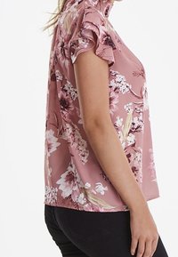 Blusa floreale rosa con maniche corte a balze, caratterizzata da un colletto alto e una vestibilità ampia. Il tessuto è leggero e ha una superficie liscia.