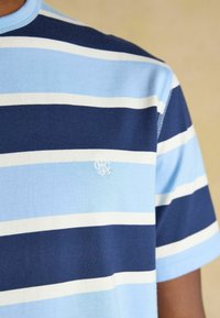 Chemise à manches courtes rayée bleu et marine avec des lignes horizontales blanches et un petit logo brodé blanc sur la poitrine.