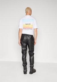 Man met kort blond haar draagt een wit grafisch t-shirt, zwarte leren broek en zwarte schoenen, staand voor een effen lichtgrijze achtergrond.