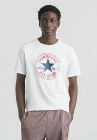 T-shirt en coton blanc avec un logo graphique rond rouge et bleu, comportant une étoile et le texte "CONVERSE CHUCK TAYLOR ALL STAR" au centre.