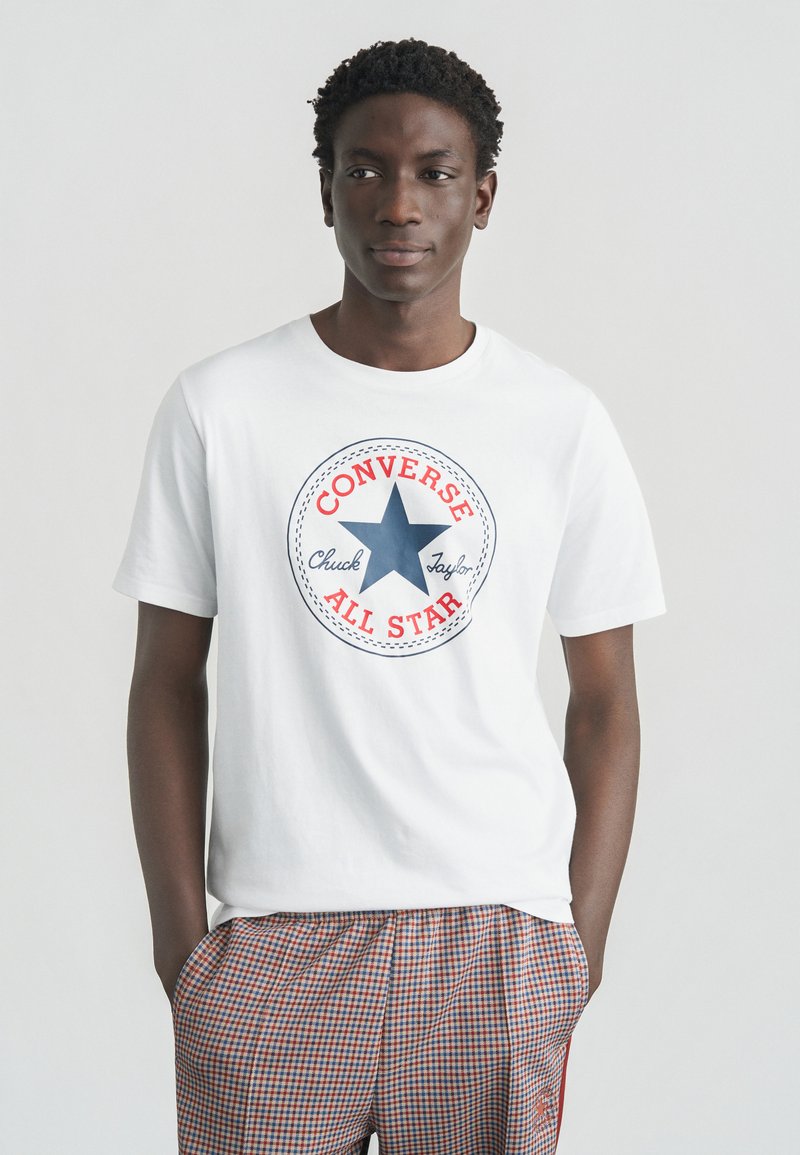 T-shirt en coton blanc avec un logo graphique rond rouge et bleu, comportant une étoile et le texte "CONVERSE CHUCK TAYLOR ALL STAR" au centre.