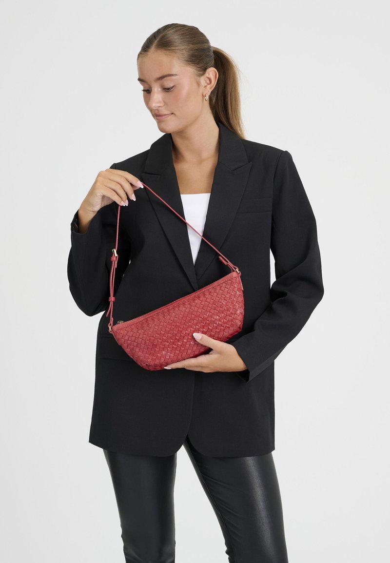 Femme en blazer noir et pantalon en cuir tenant un sac à main tissé rouge par sa bandoulière sur un fond uni.