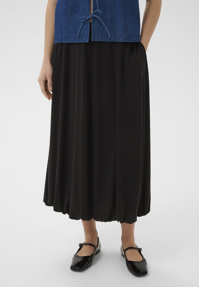 Femme portant une jupe noire ample avec un ourlet froncé, associée à des chaussures Mary Jane noires et un haut bleu avec des liens à l'avant.