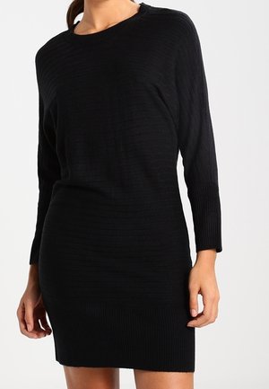 Kobieta ubrana w czarną prążkowaną dzianinową sukienkę z długim rękawem i okrągłym dekoltem, stojąca na jednolitym jasnym tle.