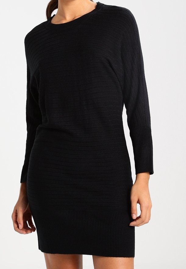 Femme portant une robe noire en maille côtelée à manches longues avec un col rond, debout devant un fond clair uni.