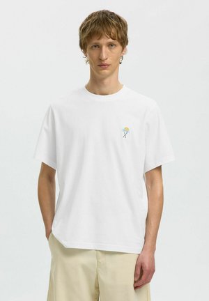 Joven con cabello castaño corto lleva una camiseta blanca con bordado pequeño de palmera y sol, pantalones cortos beige, de pie contra un fondo sencillo.