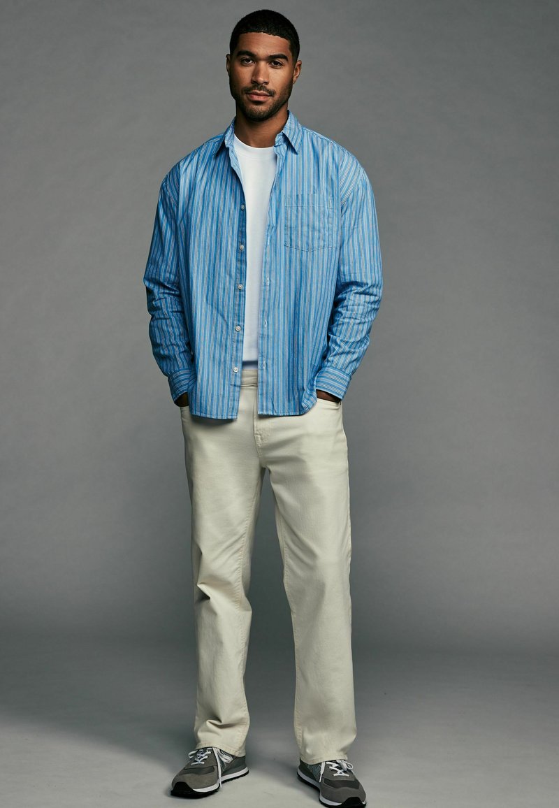 Light blue striped button-up shirt over a white t-shirt, paired with cream pants and gray sneakers. Model stands with hands in pockets.