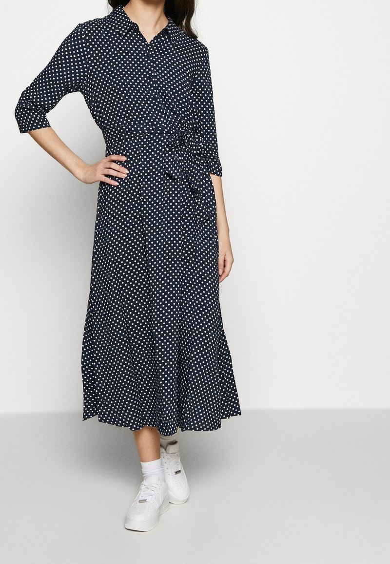 Robe bleu marine à pois blancs, boutonnée à l'avant, ceinture à la taille, manches trois-quarts, jupe fluide, portée avec des baskets blanches.