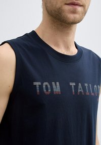 Mornarsko bez rukava majica od pamuka, s prugastim logotipom "TOM TAILOR" preko prsiju u bijeloj i crvenoj boji, s okruglim izrezom.
