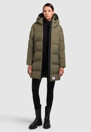 Alyvuogių žalios pūkinės striukės su kapišonu, užtrauktuko uždarymo ir juodomis detalėmis. Dėvima ant juodos bluzos ir legginsų, derinama su juodais auliniais batais.