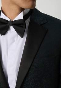 Giacca da smoking nera con motivo testurizzato blu scuro, rever a punta in satin nero, camicia bianca e papillon nero.