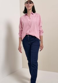 Woman wearing light pink button-up shirt, dark blue jeans, and beige cap, standing against a white and beige wall background.