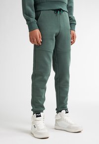 Zelené tepláky s elastickým pasem a šňůrkou, s detailem panelu. Doplňuje je bílé vysoké tenisky s logem.