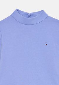 Haut col roulé bleu clair en tissu lisse, avec un petit logo rouge, blanc et bleu sur la poitrine gauche. Fermeture éclair au dos.