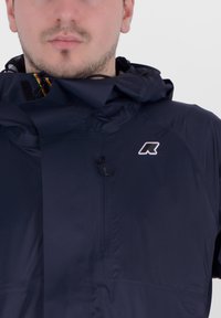 Giacca impermeabile blu navy con cappuccio, zip frontale e un piccolo logo bianco nella zona sinistra del petto, con una texture liscia.