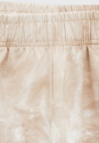 Světle béžové bavlněné tepláky s elastickým pasem a jemným vzorem tie-dye. Měkká textura s viditelnými švy podél pasu.