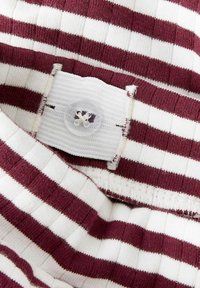 Pasiasty top w kolorach bordowym i białym, z prążkowanym wzorem i białą metką z tkaniny oraz przezroczystym zapięciem na guzik.
