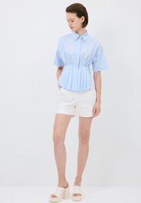 Camicetta azzurro chiaro a maniche corte con vita strutturata e design plissettato, abbinata a pantaloni corti bianchi e sandali platform cremosi e spessi.